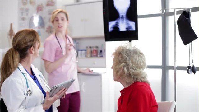 A nurse stands in the background as a doctor using an electronic tablet discusses test results with her elderly patient.
