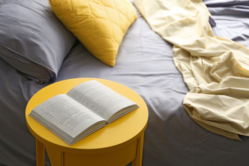 Fototapeta na wymiar Opened book on yellow bedside table near crumpled bed