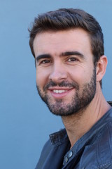 Obraz premium Handsome trendy young bearded man with a friendly smile standing against a blue background looking at the camera, head and shoulders 