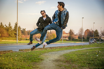 Pair of Young Men Leaping into Air and Clicking Heels Together in Unison While Walking on Path in Urban Park Alongside Paved Road at Sunset