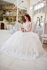 Portrait of charming red-haired bride model with wedding bouquet at hand sitting on chair at the table on great wedding hall with caves and vegetation indoor