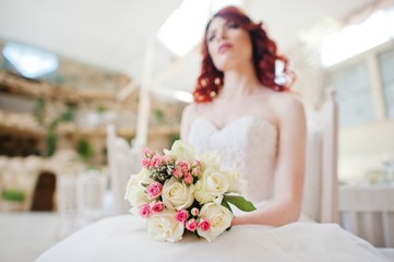 Wedding bouquet at hands of charming red-haired bride model sitting on chair at the table on great wedding hall indoor