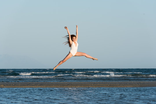 Woman with long curly hair and excellent body wear white monokini, make a dance moves at the beach. Sea and sky as background