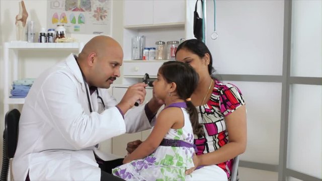 An Indian pediatrician or family doctor uses an otoscope to examine the eyes of a little girl who is being accompanied by her mother.