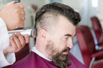 Male barber makes a mohawk hairstyle using clipper of a adult man with beard