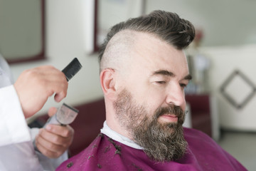 Male barber makes a mohawk hairstyle using clipper at the adult man with beard