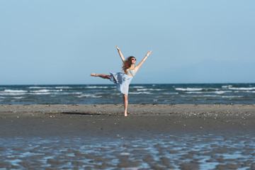 Woman with long curly hair wear dance dress and making moves, standing on the beach. Wind wave the skirt. Sea and sky as background