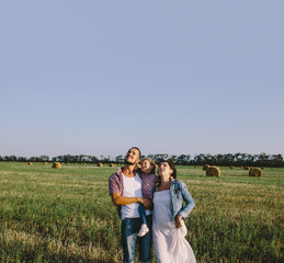 father in a plaid shirt and mother in a denim jacket and white dress hug her little daughter outdoors in field and looking up