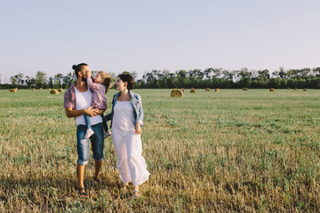 father in a red plaid shirt, daughter and pregnant mother in white dress enjoying life outdoor in field