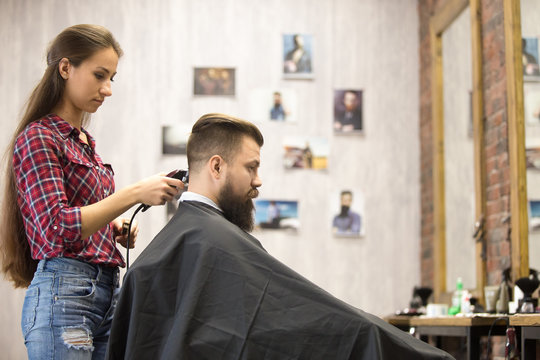 Side view portrait of handsome young bearded caucasian man getting trendy haircut in modern barbershop. Attractive woman hair stylist working, serving client, doing haircut using shaver. Indoors shot