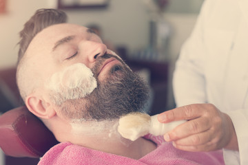 Obraz na płótnie Canvas Client with foam on face during beard shaving in barber shop. Toned