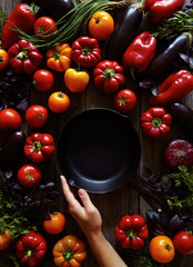 Traditional cast iron skillet pan on vintage wooden table with assorted vegetables background. Preparing ratatouille. Kitchen equipment. Top view. Hands in the frame.