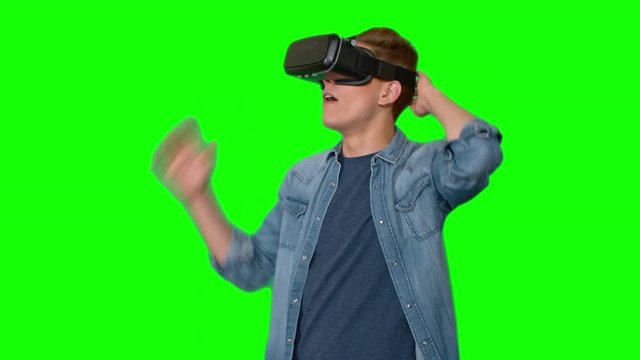 Young man with stylish haircut standing against green background putting on virtual reality goggles and looking around in amazement