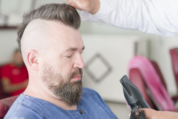 Male barber makes hair styling using a hair dryer of a adult bearded man with a mohawk