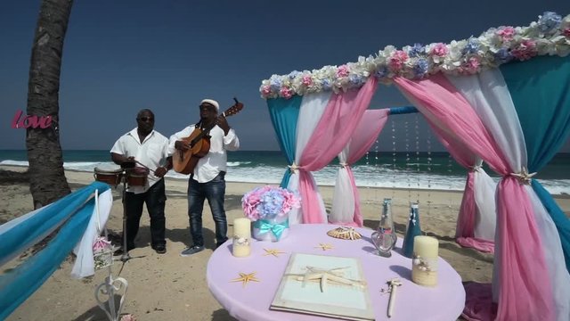 Close-up view Afro-American musicians with guitar and drum are play music, sing songs near colorful arch on the beach at a wedding ceremony in sunny day. Caribbean sea, coastline are on a background.
