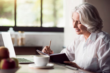 Grey-haired lady taking notes with pencil and using laptop