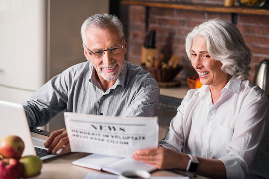 Likable Old Couple Reading Newspaper At The Kitchen Table