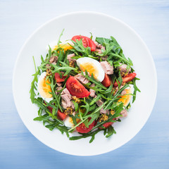 Fresh salad with tuna, tomatoes, eggs, arugula and mustard on blue wooden background top view. Healthy food.