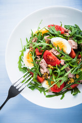 Fresh salad with tuna, tomatoes, eggs, arugula and mustard on blue wooden background top view. Healthy food.