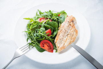Roasted chicken breast and fresh salad with tomato and greens (spinach, arugula) close up on white textured background. Healthy food.