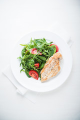 Chicken breast and fresh salad with tomato and greens (spinach, arugula) top view on white wooden background. Healthy food.