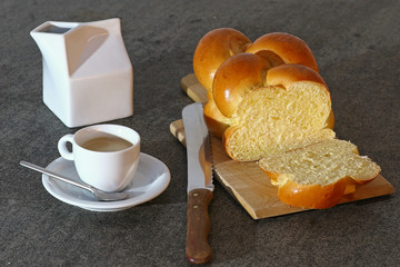 Fresh braided bread with knife on a wooden cutting board, espresso coffee cup and milk or cream carton