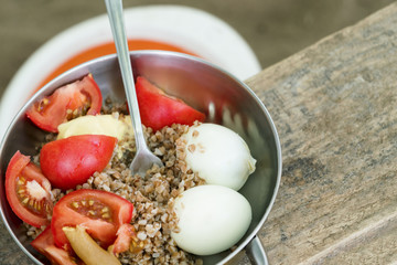 Iron bowl with buckwheat, tomatoes, boiled egg and fork on a wooden table