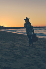 Silhouette of elegant beautiful female having fun playing guitar on sand beach outside sunset sky background