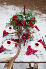 Wedding table setting in marsala colors with plates, cutlery, red floral compositions, candles, velvet napkins, deer horns on table covered with snow