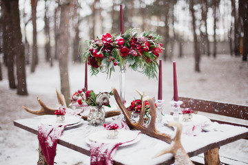 Wedding table setting in marsala colors with plates, cutlery, red floral compositions, candles, velvet napkins, deer horns on table covered with snow