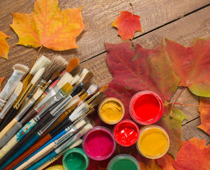 Obraz premium Colorful paints, brushes, palette, autumn leaves on wood background. Autumn still-life. The workplace of the artist. Banner for the school.