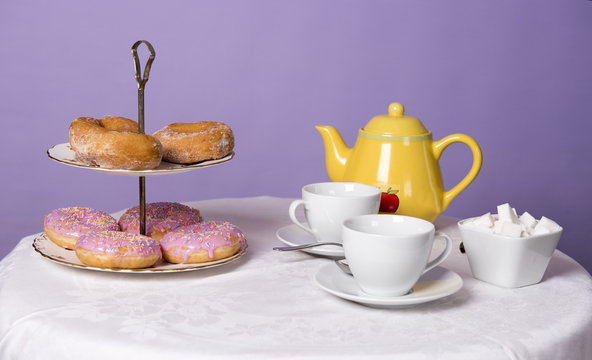 Donuts on a stand with a pot of tea and tea cups on a white table. 