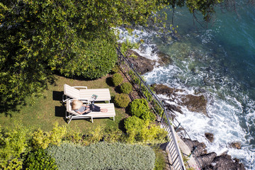 High angle view shot of a woman lying on lounge chair and using her laptop while relaxing by the sea. 