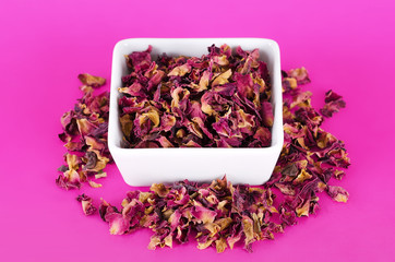 Obraz na płótnie Canvas Rose petals in a white bowl on pink background. Dried blossoms, used for perfumes, cosmetics, teas and baths. Purple and orange colored organic herb. Isolated macro photo close up from above.