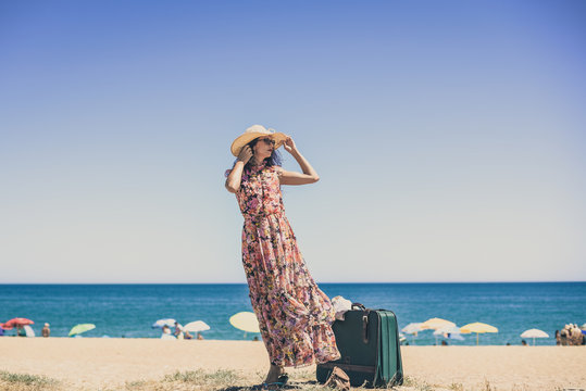 Beautiful elegant female carrying suitcase walking on sandy beach blue sky background - Powered by Adobe