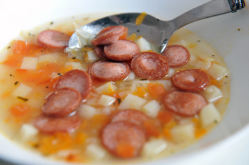Vegetable soup with added pieces of frankfurter sausages in a white plate.