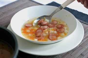 Vegetable soup with added pieces of frankfurter sausages in a white plate.