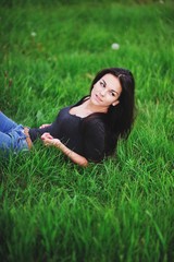Charming young long haired brunette girl in black blouse poses for the camera on the grass, looking to the side.
