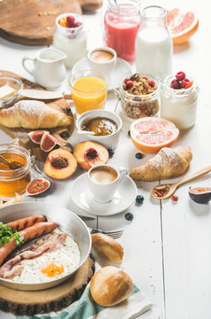 Fried egg with sausages and bacon, bread, croissants with jam and butter, fruits, smoothie, orange juice, yogurt, granola with milk and coffee on white wooden background. Selective focus, copy space