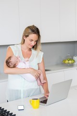 Mother using laptop while carrying baby in kitchen