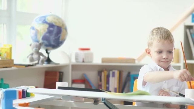 Interior of a child's room. Globe and toys on the background. Boy paints watercolors. Boy 5-7 years old sitting at a desk and draws. A boy holding a brush to paint.