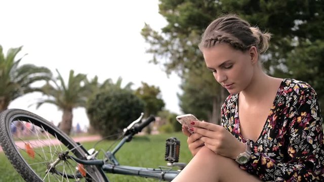 Pretty cute girl using smartphone beside her bike in the park with palms on a sunny day