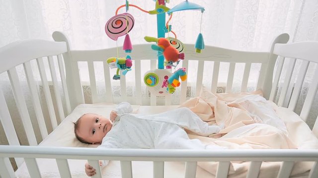 Cute 3 months old baby lying in white wooden crib next to big window