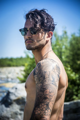 Athletic shirtless young man outdoor at river or water stream, looking away, with rocks and stones in background