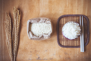 Cooked rice in bowl with raw rice grain and dry rice plant on wooden table background.