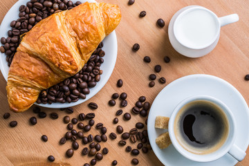 Breakfast coffee - cup, sugar, milk jug and croissant with coffee beans on wooden background.