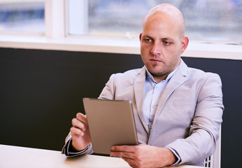 Business man holding and using a high tech tablet while seated at a conference table early in the morning with a window behind him and light coming in through the glass.
