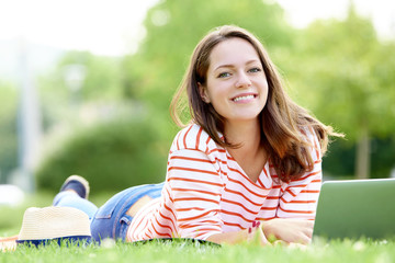Enjoy summer sunshine at park. Full length portrait of young woman with her laptop lying on grass outdoor.
