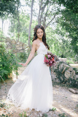 Fototapeta na wymiar portrait of a beautiful happy brunette bride in wedding white dress holding hands in pink and purple bouquet of flowers outdoors on the background of green leaves