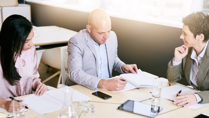 Business meeting between three executive employees, two female and one male, taking place in a birght conference table early in the morning to prepare for the week ahead.
